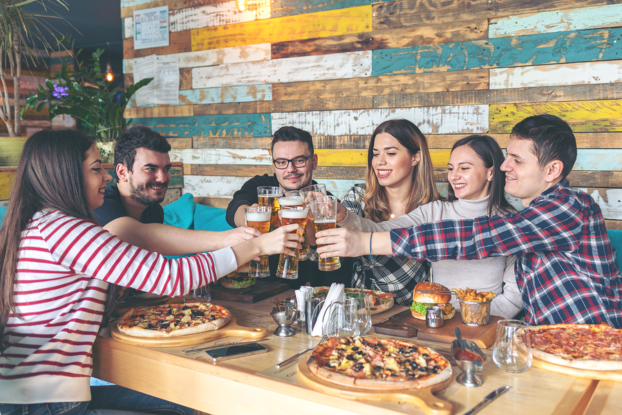 Happy young hipster friends celebrating by toasting with beer eating pizza and burgers at bar restaurant - Friendship concept with young people enjoying time together and having genuine fun at rustic pizzeria - cheers
