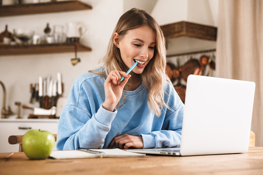 Portrait of caucasian blond woman 20s wearing casual sweatshirt working on laptop and writing down notes while sitting at wooden table at home