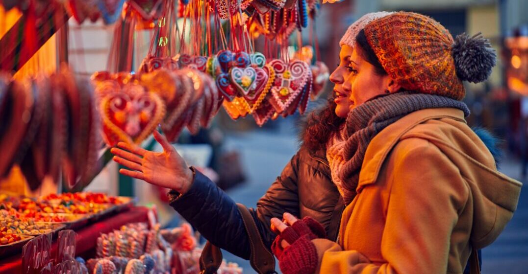 2 Freunde an einem Weihnachtsmarktstand unterhalten sich über einzigartige Weihnachtstraditionen