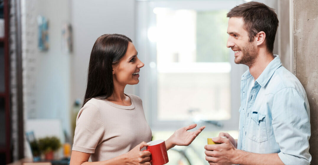 break. Two cheerful young people holding coffee cups and talking while standing in office