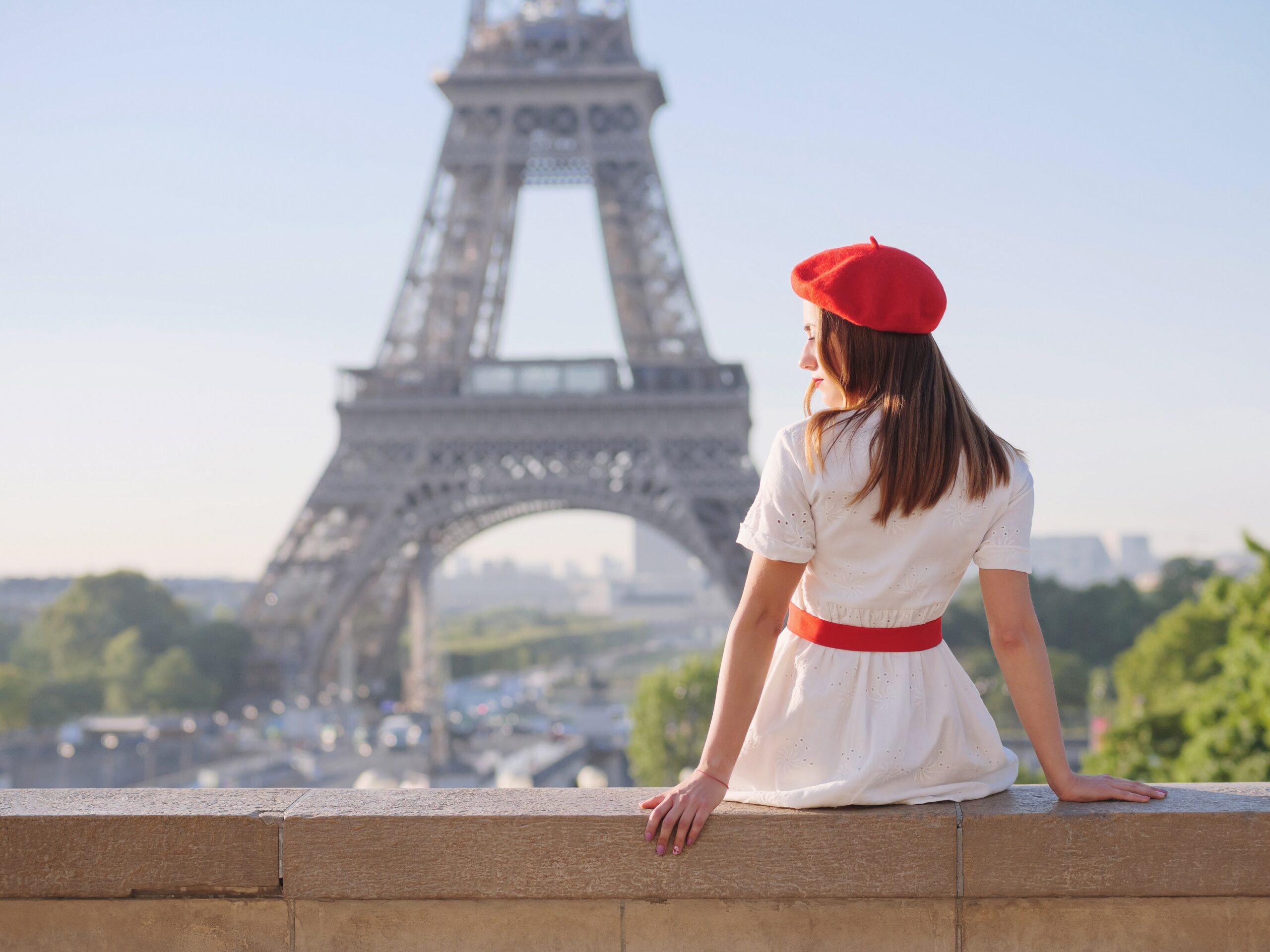 A girl with a beret sitting on a wall in front of the Eiffel power like Emily in paris