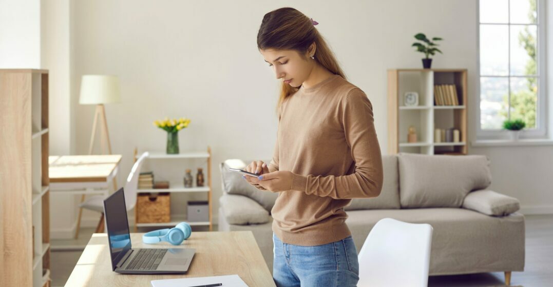 A woman standing with her phone in front of her laptop looking up the best German online dictionaries
