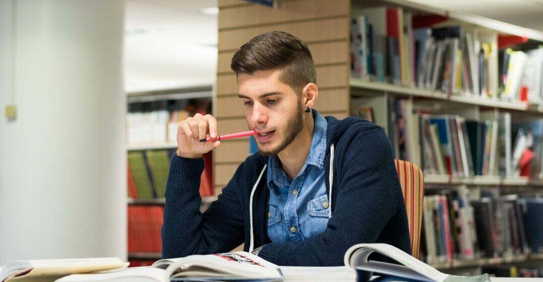 Young man with books in a library studying a list of English prepositions