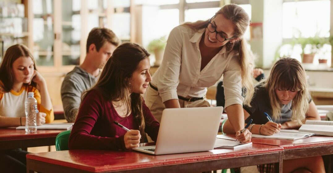 Smiling woman who is teaching English as a second language explains something to her student with a laptop in a classroom