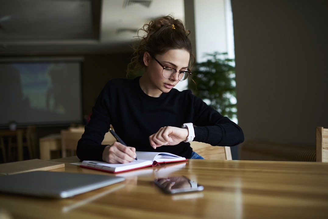 Young woman writing into a notebook looking at her watch to study expressions for time