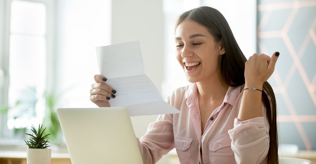 Junge Frau an ihrem Laptop hält ein Motivationsschreiben auf Englisch in der Hand und freut sich darüber