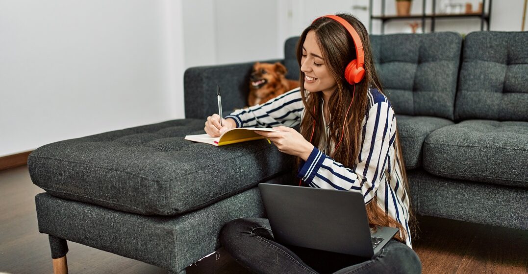 Young woman with a laptop sitting on the floor writing down spanish past tense conjugations in a notebook
