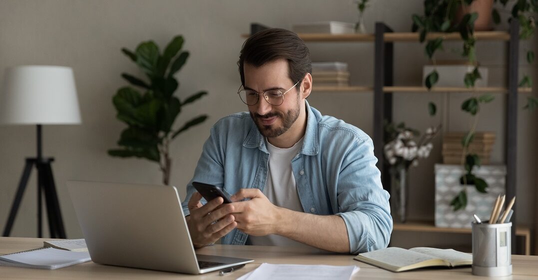 Man sitting at his laptop holding his phone studying with an English pronoun chart