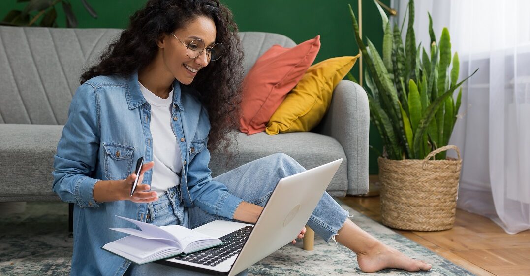 Young woman with curls sitting on the floor with her laptop smiling and studying the reflexive verbs in Spanish