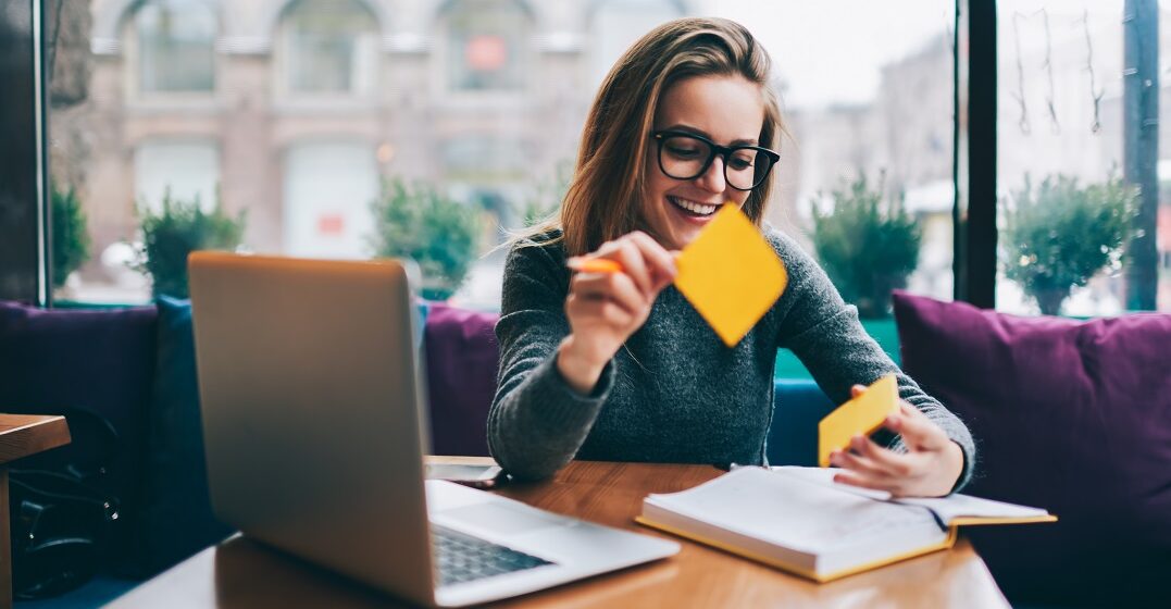 Eine junge Frau vor ihrem Laptop hält Post-its in der Hand, mit denen sie deutsche Abkürzungen lernt