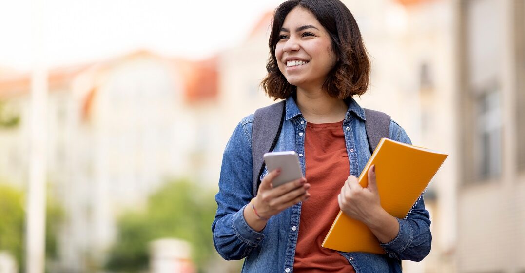 Eine lächelne junge Frau hält ein Buch und ihr Smartphone und überlegt, was die Unterschiede ziwschen wenn und als sind