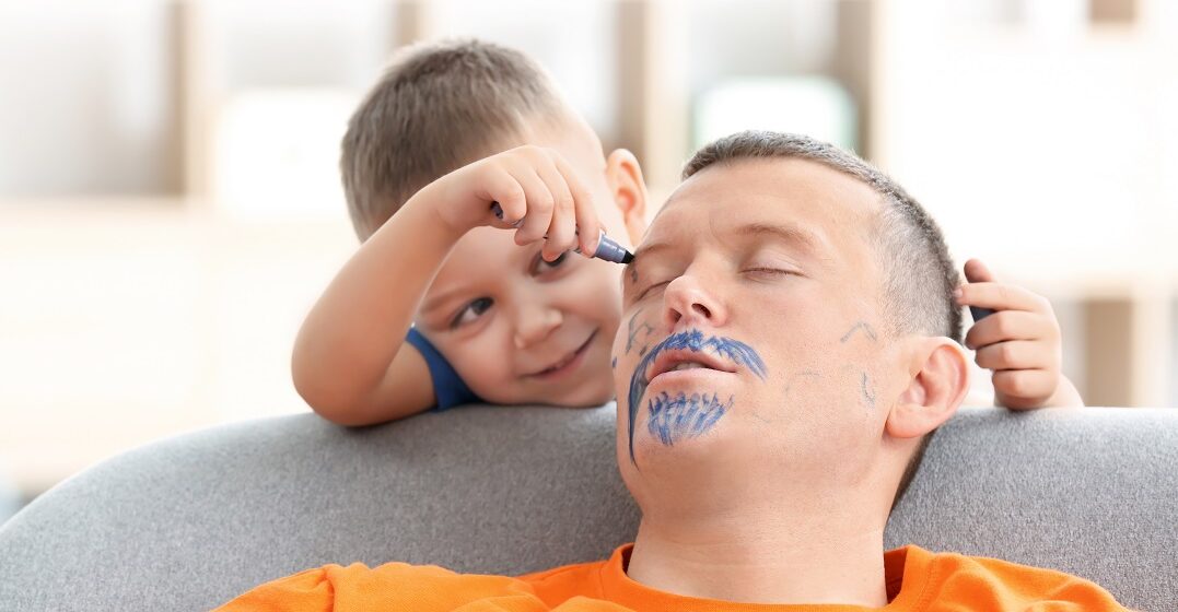 Little boy painting his father's face while he sleeping. April fool's day prank