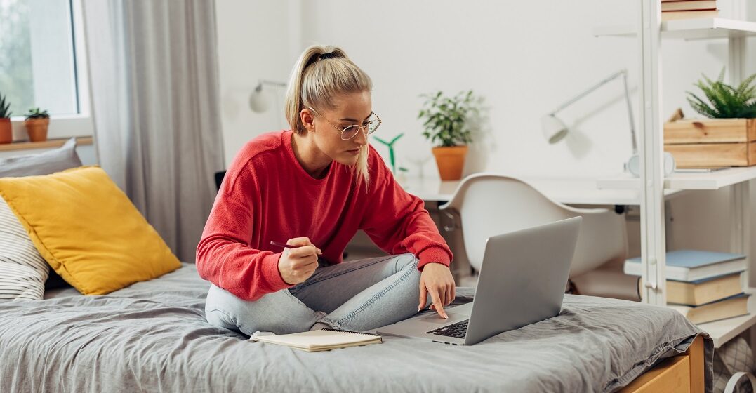 Eine junge Frau sitzt auf ihrem Bett am Laptop und lernt online über den Infinitiv mit zu auf Deutsch