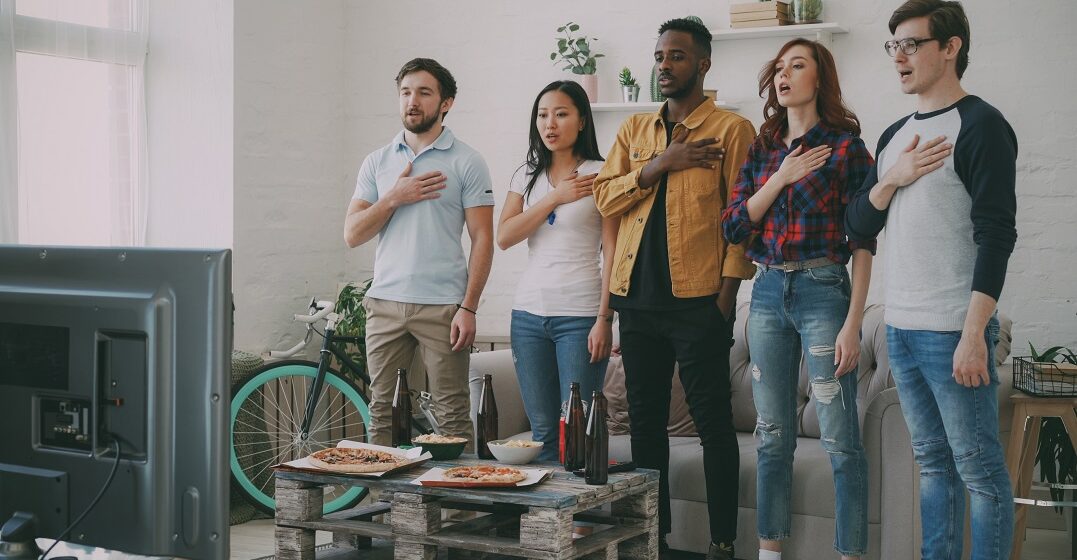 Multiethnic group singing the Swiss national anthem in front of a TV