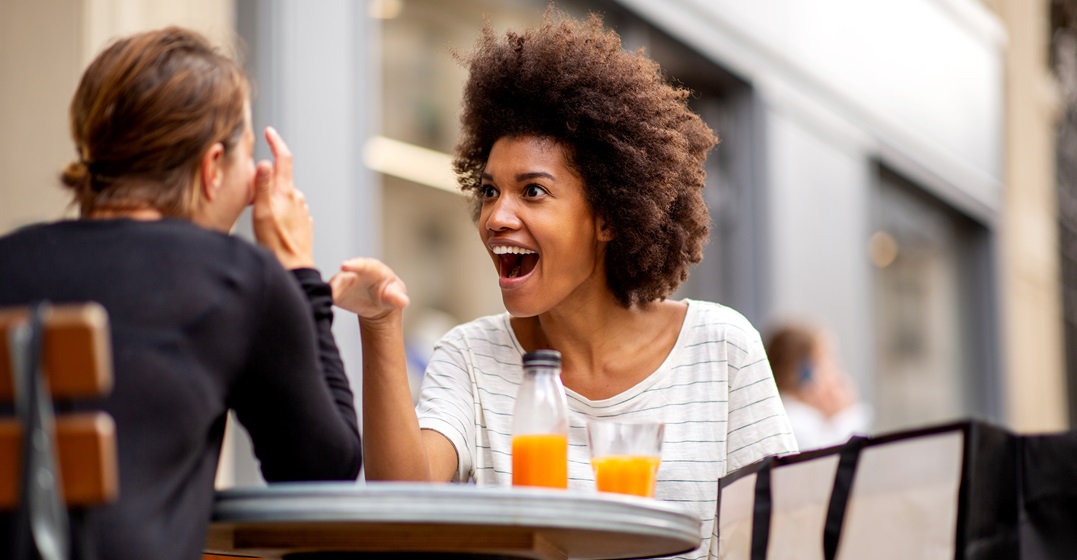 Two young women sitting outside of a café, one looks surprised and uses German interjections