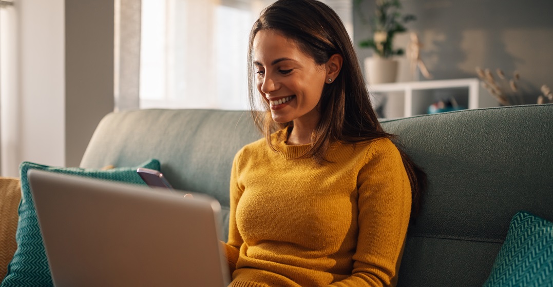 Cheerful woman sitting on her couch with a laptop studying the most essential Spanish abbreviations