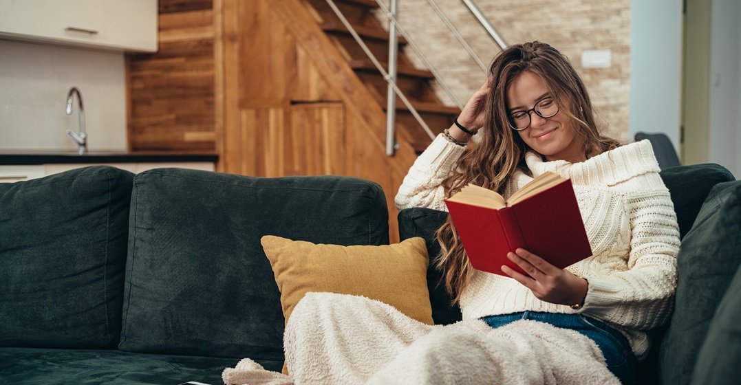 Junge Frau auf einem Sofa sitzt mit einer Decke und einem Buch und liest deutsche Märchen