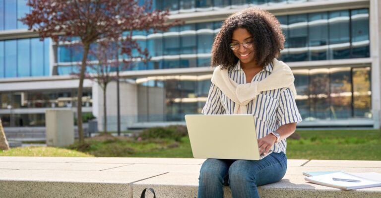 Junge afrikanische Frau sitzt vor einem Gebäude an ihrem Laptop und macht einen Online_sprachkurs
