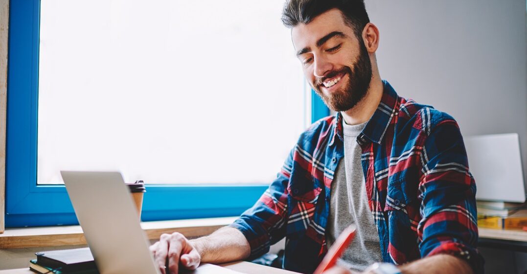 smiley young man sitting at the table with his laptop while writing down on a notebook the days of the week in Italian