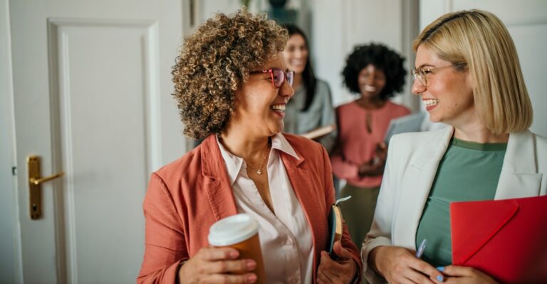 Zwei Frauen unterhalten sich lachend bei der Arbeit, einen Kaffee und eine Mappe haltend.