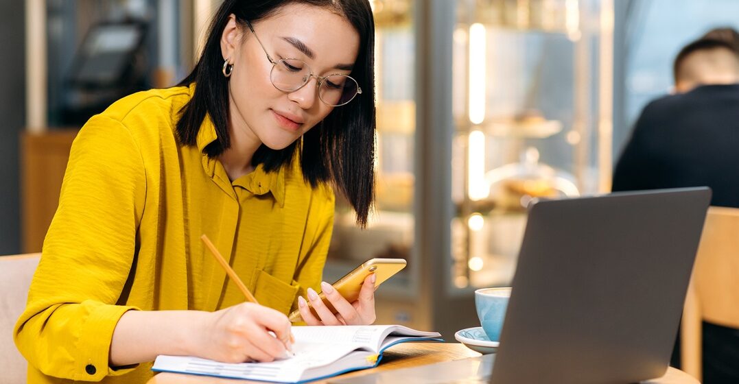 Asian girl on her laptop and writing down on her notebook notes about the fare conjugation in italian