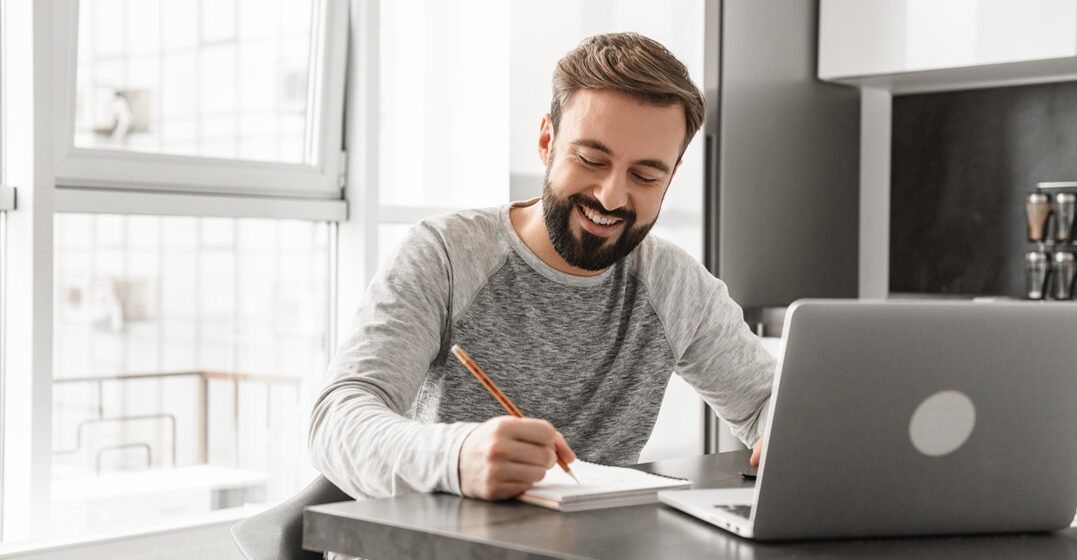 young man smiling and sitting at his desk learning italian grammar and taking notes on a notepad