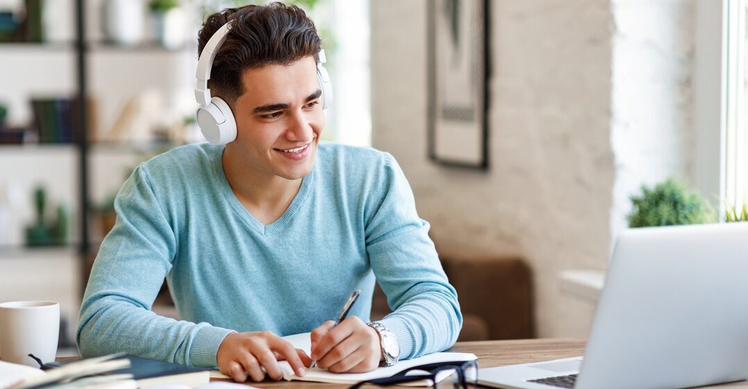 young caucasian man, sitting at his desk with headphones on and writing down notes on italian verbs