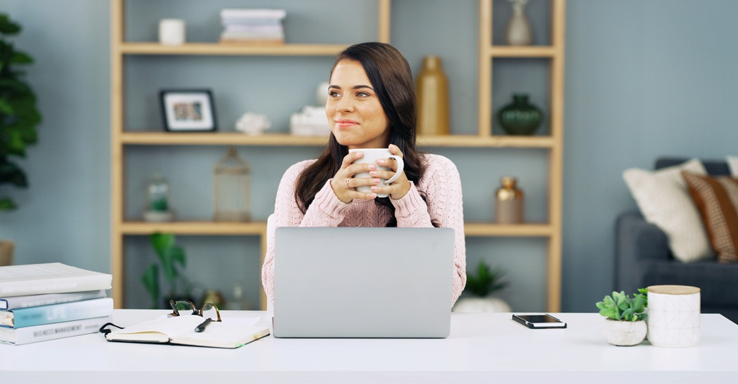 Eine Frau hält eine Kaffeetasse und sitzt vor ihrem Laptop und überlegt, wie man eine E-Mail auf Deutsch schreibt