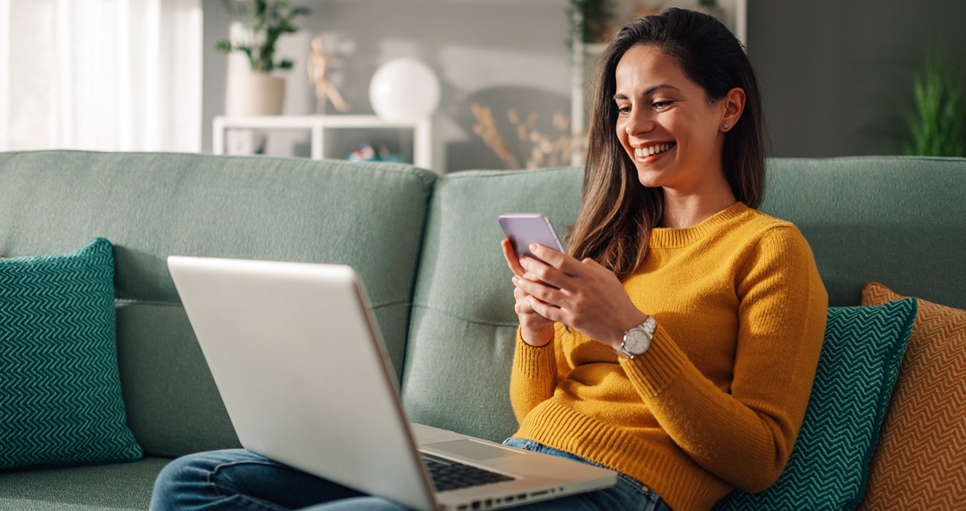 Eine lächelnde Frau sitzt auf dem Sofa an ihrem Laptop und hält ihr Smartphone dabei und übt die Temporaladverbien