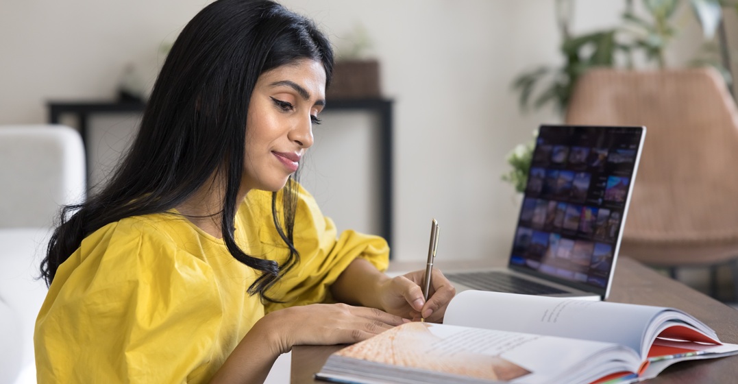 Eine Frau schreibt in ein Lehrbuch und hat dabei ihren Laptop offen, sie lernt die Benutzung des Apostroph im Deutschen