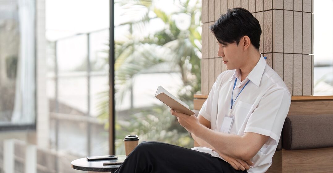 Young Asian man, sitting in a well-lit room, completing his German reading practice with a double translated book.