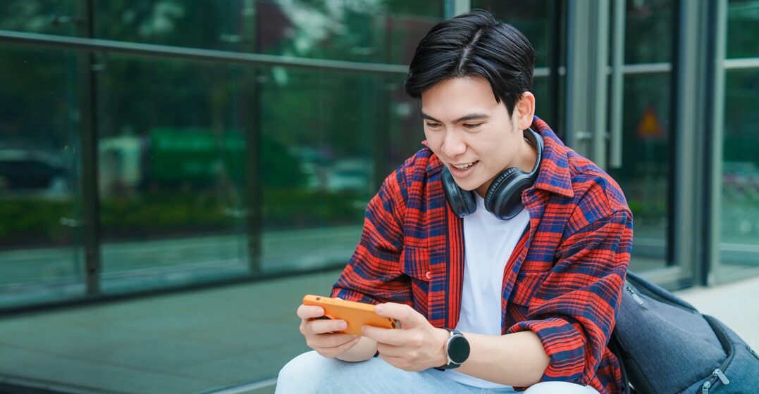 young asian man sitting with his phone playing some games in german language to boost his learning