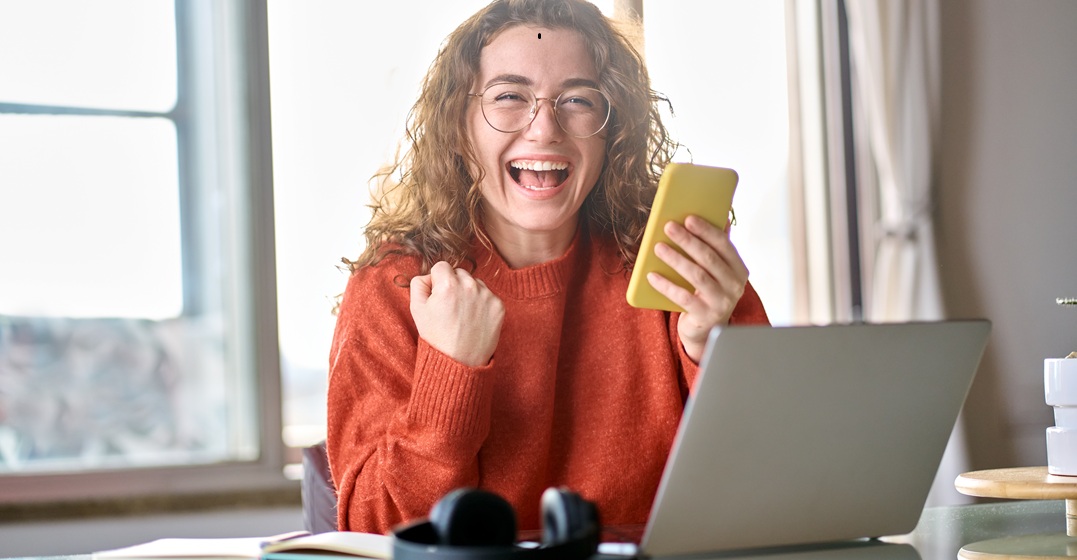 Happy young woman holding her phone, sitting at her laptop smiling because she won the Lingoda scholarship
