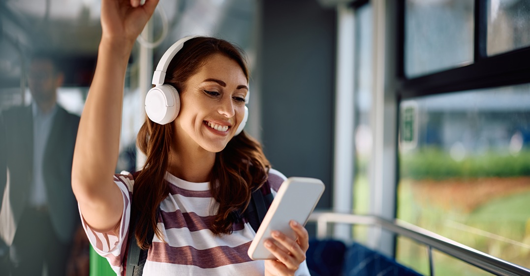 Eine lächelnde Frau steht in der Straßenbahn und hält ihr SMartphone und lernt dabei Deutsch mit Musik über Kopfhörer