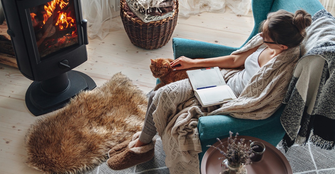 Young woman practicing German Gemütlichkeit in an arm chair with a book and her cat in front of a fireplace